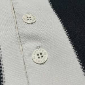 Puede incluir: Primer plano de un polo a rayas blancas y negras. La camisa tiene tres botones blancos con el logotipo de Adidas. La tela tiene una apariencia texturizada y las rayas son verticales.
