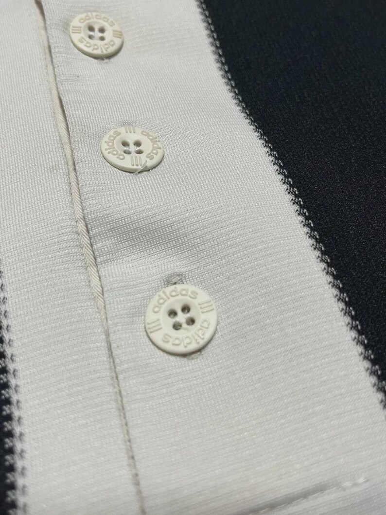 Puede incluir: Primer plano de un polo a rayas blancas y negras. La camisa tiene tres botones blancos con el logotipo de Adidas. La tela tiene una apariencia texturizada y las rayas son verticales.