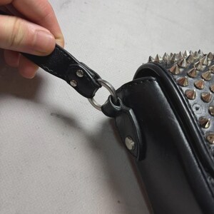 Puede incluir: Bolso de mano de cuero negro con tachuelas de metal plateado y una correa. El bolso tiene un cierre de solapa y un anillo de metal plateado que conecta la correa al bolso.