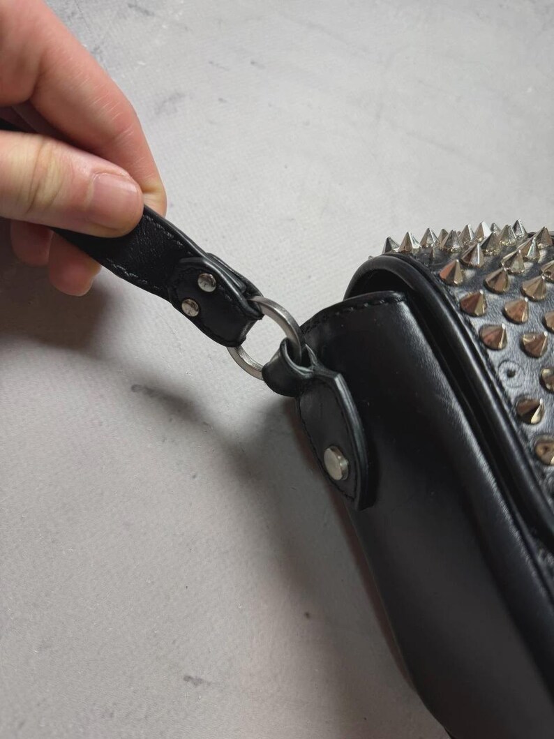 Puede incluir: Bolso de mano de cuero negro con tachuelas de metal plateado y una correa. El bolso tiene un cierre de solapa y un anillo de metal plateado que conecta la correa al bolso.