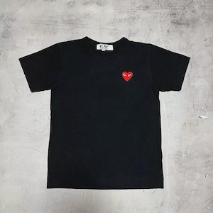 Puede incluir: Camiseta negra de manga corta con un pequeño gráfico en forma de corazón rojo en el pecho. La camiseta tiene cuello redondo y la palabra "PLAY" está impresa en la parte superior izquierda.