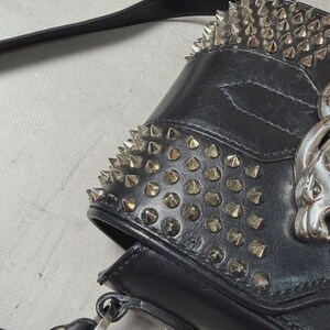 Puede incluir: Bolso de hombro de cuero negro con tachuelas c&oacute;nicas plateadas en la parte superior y la solapa delantera. El bolso presenta un emblema de calavera plateada y una correa larga y ajustable. El bolso tiene una est&eacute;tica g&oacute;tica o punk.