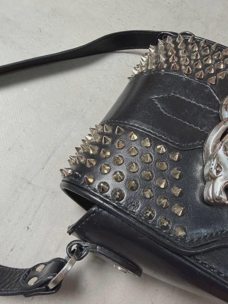 Puede incluir: Bolso de hombro de cuero negro con tachuelas c&oacute;nicas plateadas en la parte superior y la solapa delantera. El bolso presenta un emblema de calavera plateada y una correa larga y ajustable. El bolso tiene una est&eacute;tica g&oacute;tica o punk.