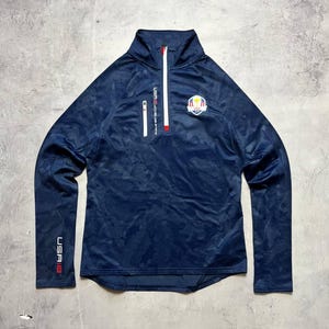 Könnte beinhalten: Marineblaues Langarm-Sportoberteil mit Camouflage-Muster. Mit weißem Reißverschluss, kleiner Brusttasche und Logo-Patch. Am Ärmelaufschlag steht der Text "USA" in Rot.