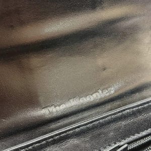 Puede incluir: Primer plano del interior de una cartera de cuero negro. El texto en relieve "The Kooples" es visible. La cartera tiene una cremallera y una superficie texturizada.