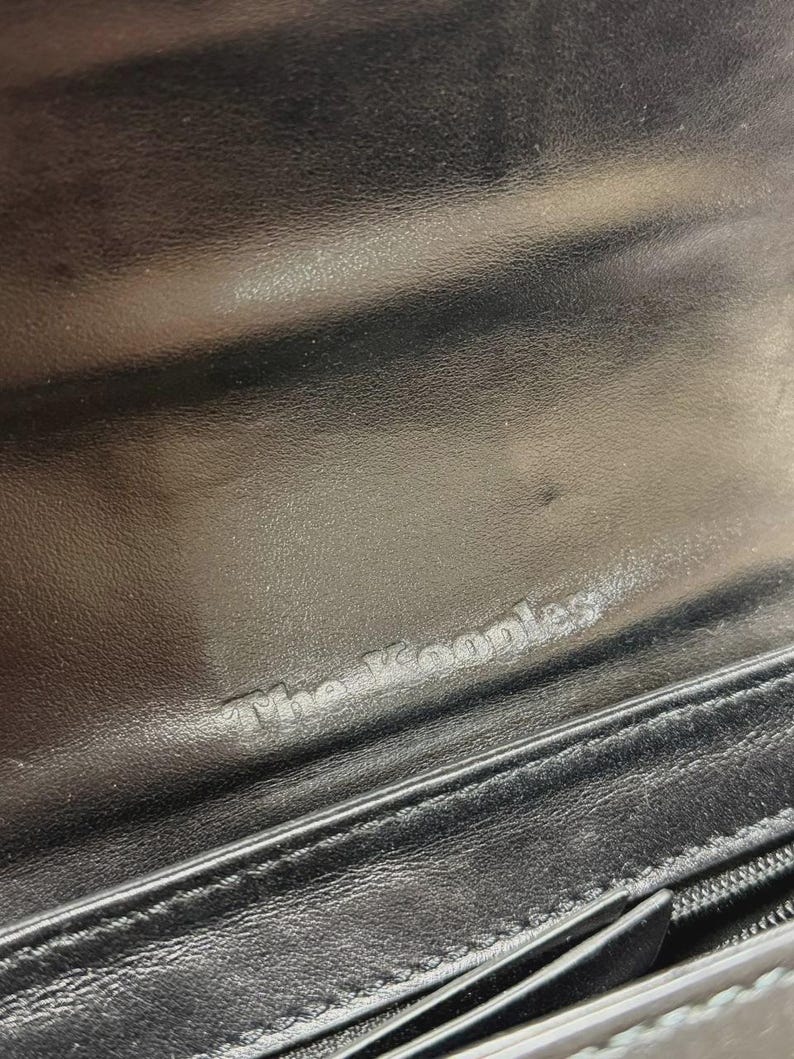 Puede incluir: Primer plano del interior de una cartera de cuero negro. El texto en relieve "The Kooples" es visible. La cartera tiene una cremallera y una superficie texturizada.