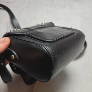 Puede incluir: Bolso bandolera de cuero negro con tachuelas de metal plateado en la solapa. El bolso tiene forma rectangular con base redondeada y una correa larga y ajustable. El bolso es de cuero liso y brillante.