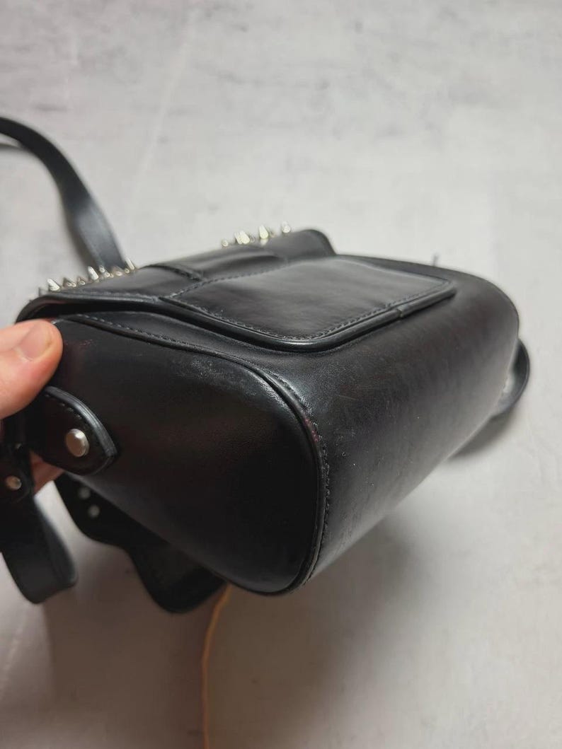 Puede incluir: Bolso bandolera de cuero negro con tachuelas de metal plateado en la solapa. El bolso tiene forma rectangular con base redondeada y una correa larga y ajustable. El bolso es de cuero liso y brillante.