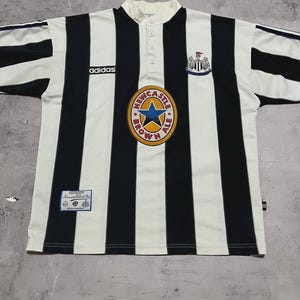 Puede incluir: Una camiseta de f&uacute;tbol vintage del Newcastle United con rayas verticales negras y blancas. Presenta el logotipo de Adidas, un escudo de Newcastle Brown Ale y el escudo del club. La camiseta tiene cuello abotonado y mangas cortas.