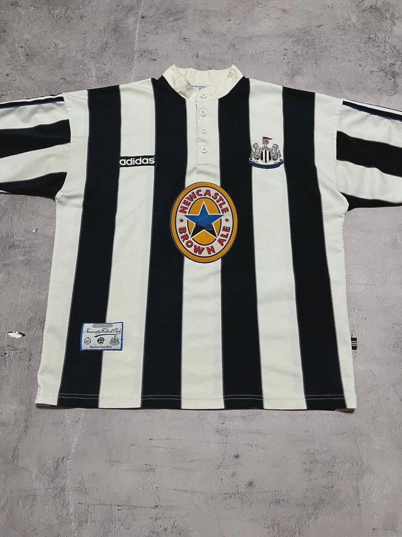 Puede incluir: Una camiseta de f&uacute;tbol vintage del Newcastle United con rayas verticales negras y blancas. Presenta el logotipo de Adidas, un escudo de Newcastle Brown Ale y el escudo del club. La camiseta tiene cuello abotonado y mangas cortas.