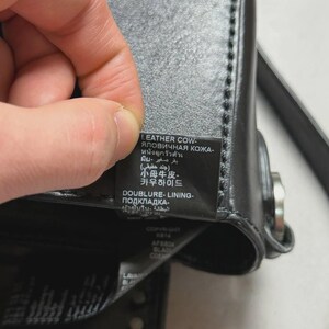 Puede incluir: Primer plano de un bolso de cuero negro con una etiqueta. La etiqueta incluye texto en varios idiomas, incluyendo ingl&eacute;s, ruso, tailand&eacute;s, &aacute;rabe, coreano y japon&eacute;s. El bolso tiene un cierre plateado y tachuelas decorativas.