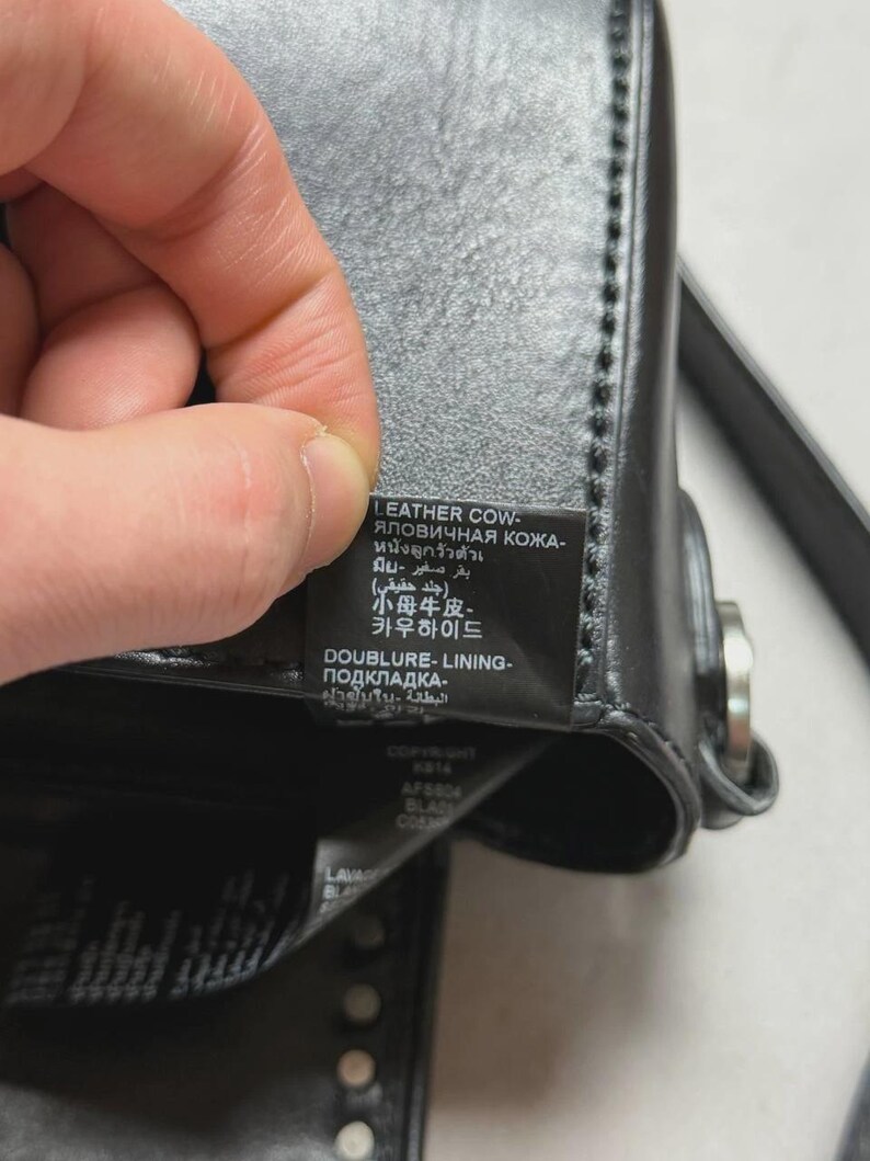 Puede incluir: Primer plano de un bolso de cuero negro con una etiqueta. La etiqueta incluye texto en varios idiomas, incluyendo ingl&eacute;s, ruso, tailand&eacute;s, &aacute;rabe, coreano y japon&eacute;s. El bolso tiene un cierre plateado y tachuelas decorativas.