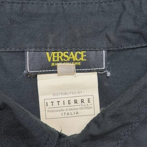 May include: Close-up of a dark gray button-down shirt with the Versace Jeans Couture label. The label is yellow with black text. A white tag below the label reads "Distributed by: ITTIERRE" and other text. The shirt is buttoned at the collar.