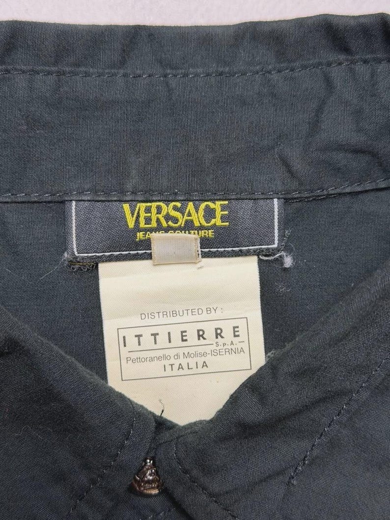 May include: Close-up of a dark gray button-down shirt with the Versace Jeans Couture label. The label is yellow with black text. A white tag below the label reads "Distributed by: ITTIERRE" and other text. The shirt is buttoned at the collar.