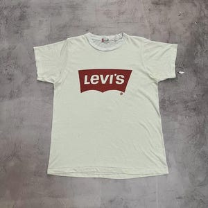 Pode incluir: Camiseta creme com o logotipo da Levi's em um retângulo vermelho. O logotipo está centralizado no peito. A camiseta tem mangas curtas e gola redonda. O tecido parece ser algodão macio e desgastado.