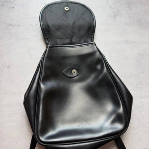 May include: Black backpack with a flap closure and adjustable straps. The bag is made of a smooth, shiny material and has a small button closure on the front. The interior lining is black.