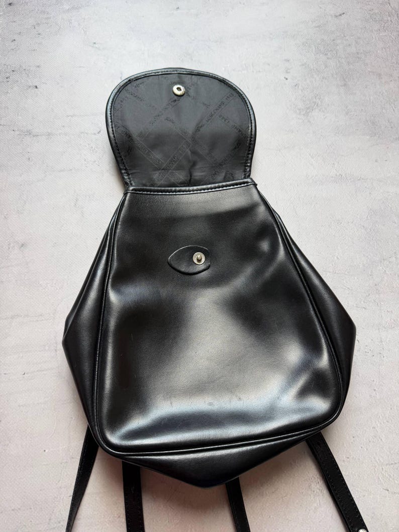 May include: Black backpack with a flap closure and adjustable straps. The bag is made of a smooth, shiny material and has a small button closure on the front. The interior lining is black.