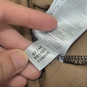 Puede incluir: Primer plano de una etiqueta blanca con los n&uacute;meros "07/15", "F81861" y "11N004" impresos en negro. La etiqueta est&aacute; adherida a una tela marr&oacute;n. La tela tiene un aspecto texturizado.