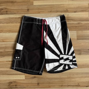 Pode incluir: Calções de banho pretos e brancos com um padrão de raios de sol na perna direita e a perna esquerda preta. Os calções incluem um bolso lateral branco com dois ilhós, um cordão vermelho na cintura e um emblema retangular da marca Billabong.