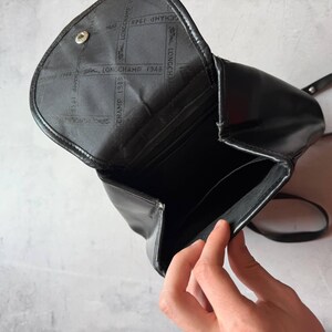 May include: Black leather backpack with a flap closure. The interior is visible, revealing pockets and the Longchamp 1948 logo. The bag has a single strap and a silver-tone button closure. The bag is open, revealing the interior.