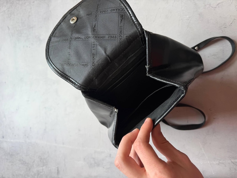 May include: Black leather backpack with a flap closure. The interior is visible, revealing pockets and the Longchamp 1948 logo. The bag has a single strap and a silver-tone button closure. The bag is open, revealing the interior.