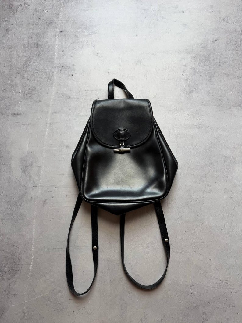 May include: Black leather backpack with a flap closure and silver-tone hardware. The bag has adjustable shoulder straps and a top handle. The backpack is shown against a gray background.