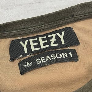 Puede incluir: Primer plano de una camiseta color beige con cuello y ribetes verde oscuro. Dos etiquetas de tela negra est&aacute;n cosidas a la camiseta. La etiqueta superior dice "YEEZY" en letras blancas. La etiqueta inferior dice "SEASON 1" con el logo de Adidas.
