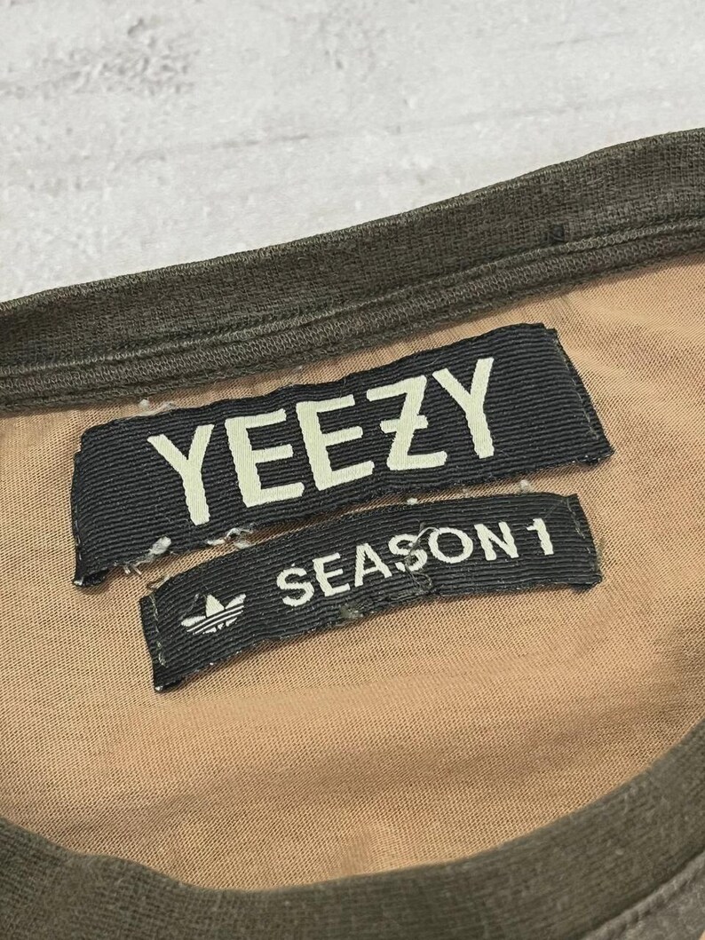 Puede incluir: Primer plano de una camiseta color beige con cuello y ribetes verde oscuro. Dos etiquetas de tela negra est&aacute;n cosidas a la camiseta. La etiqueta superior dice "YEEZY" en letras blancas. La etiqueta inferior dice "SEASON 1" con el logo de Adidas.