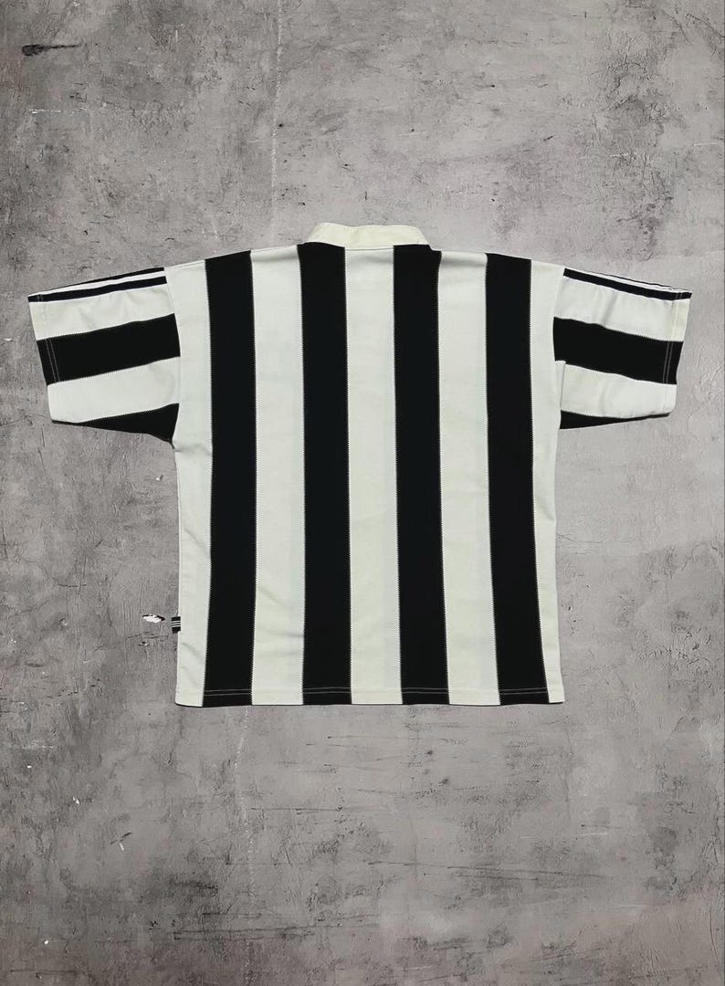 Puede incluir: Camiseta de manga corta a rayas blancas y negras. La camiseta tiene cuello blanco y rayas negras verticales en la parte delantera y trasera. Las mangas tambi&eacute;n tienen rayas blancas y negras. La camiseta es de material ligero.