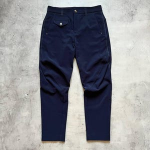 May include: Dark navy trousers with a tailored design. They have a button fastening, a small pocket with a button, and delicate stitching. The fabric has a subtle sheen, and the trousers seem to be made of a lightweight material.