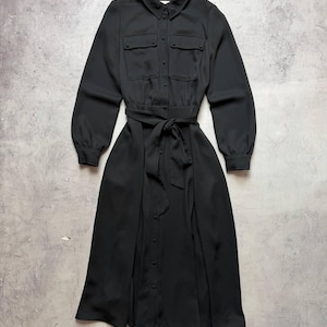 May include: Black long-sleeve dress with a collared neckline and button-down front. The dress features two chest pockets, a belted waist, and a flowing skirt. The dress is made of a smooth, solid black fabric.