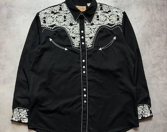 May include: A black long-sleeve button-down shirt featuring white floral embroidery across the shoulders and cuffs. The shirt includes a pointed collar, a front placket with metallic snap buttons, and decorative arrow-shaped stitching details.