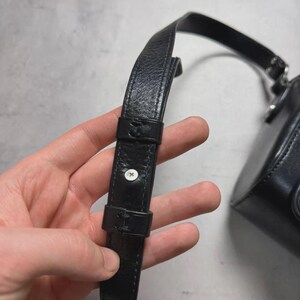 Puede incluir: Primer plano de una correa de bolso de cuero negro con una hebilla y un bot&oacute;n plateado. La correa est&aacute; sujeta a un peque&ntilde;o bolso redondo de cuero negro. El fondo es una superficie gris claro.