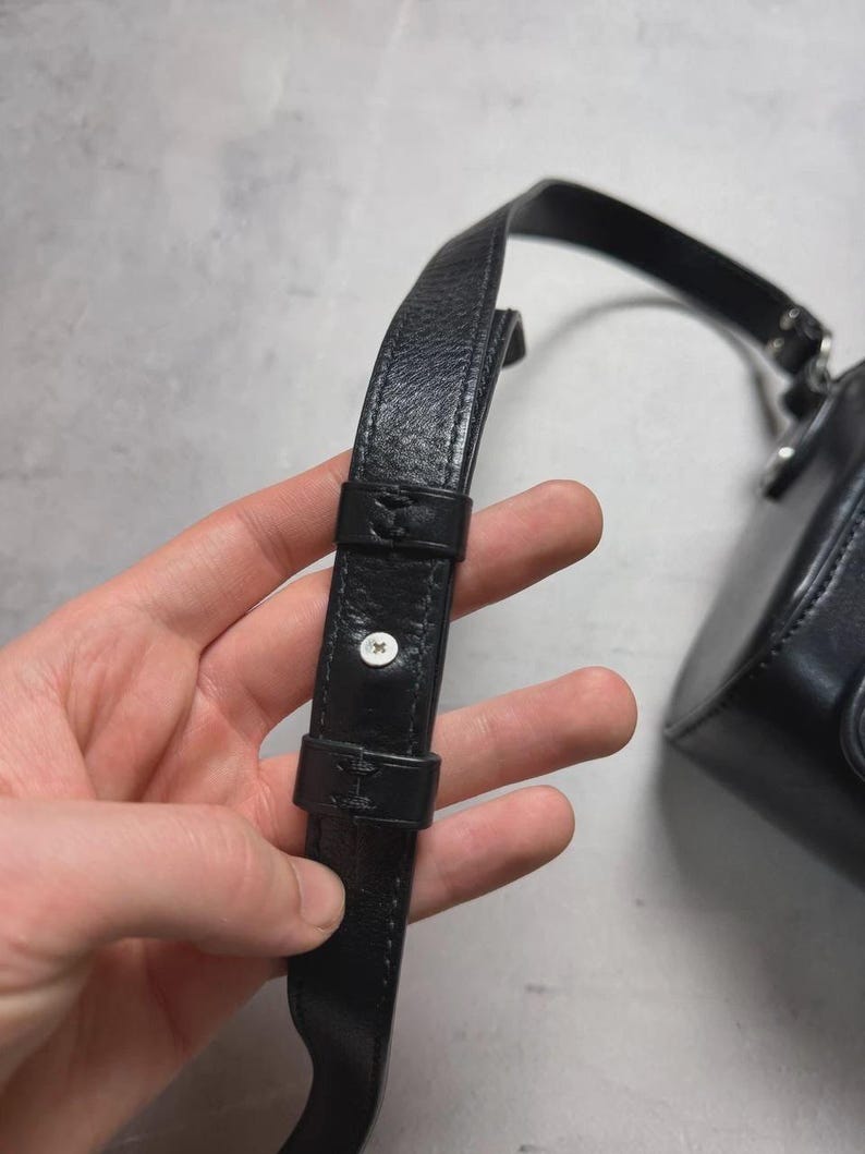 Puede incluir: Primer plano de una correa de bolso de cuero negro con una hebilla y un bot&oacute;n plateado. La correa est&aacute; sujeta a un peque&ntilde;o bolso redondo de cuero negro. El fondo es una superficie gris claro.