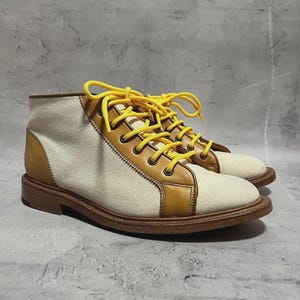 May include: A pair of canvas and leather ankle boots. The boots feature a cream-colored canvas upper with tan leather accents and a brown sole. Bright yellow laces add a pop of color. The boots are designed for everyday wear.