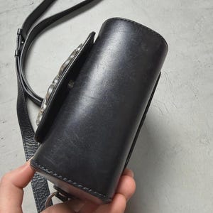 Puede incluir: Bolso bandolera de cuero negro con cierre plateado. El bolso tiene forma cil&iacute;ndrica y una correa larga y ajustable. El bolso est&aacute; sujeto por una mano.