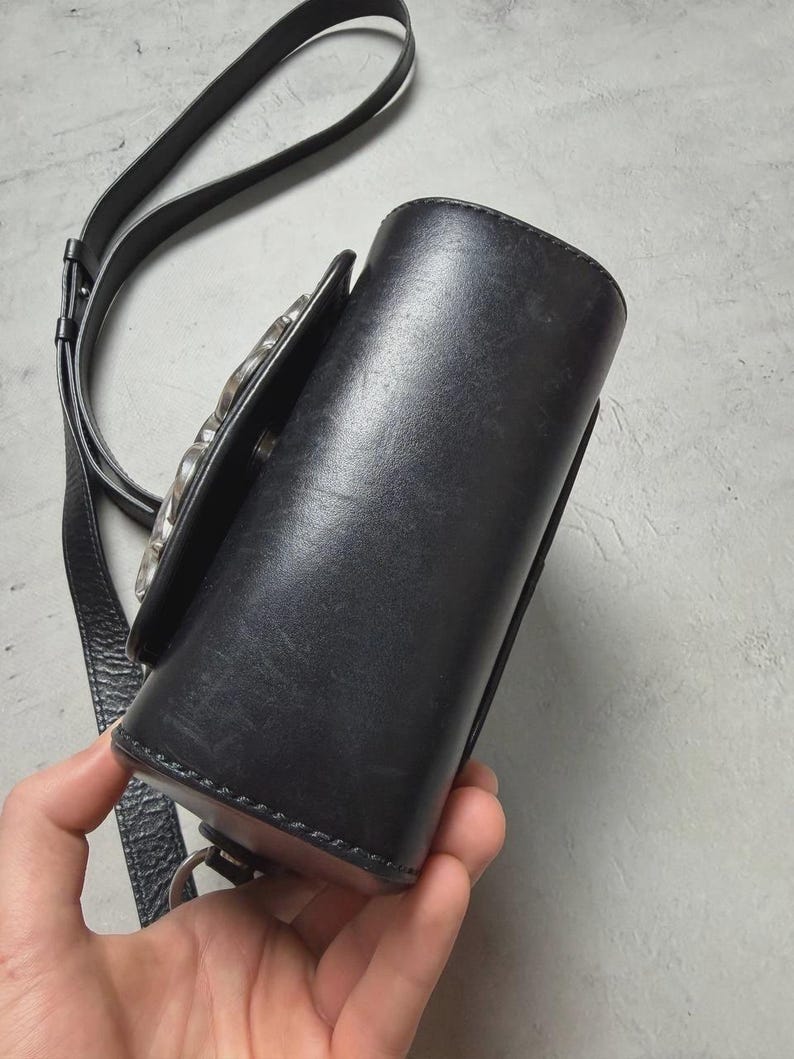 Puede incluir: Bolso bandolera de cuero negro con cierre plateado. El bolso tiene forma cil&iacute;ndrica y una correa larga y ajustable. El bolso est&aacute; sujeto por una mano.