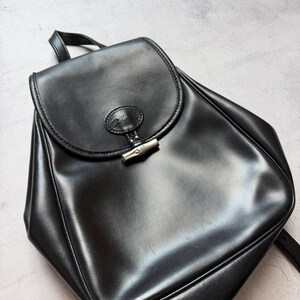 May include: A black leather backpack with a flap closure and a silver-tone clasp. The bag has a top handle and adjustable shoulder straps. The leather has a glossy finish, and the bag appears to be in good condition. The overall design is simple and classic.