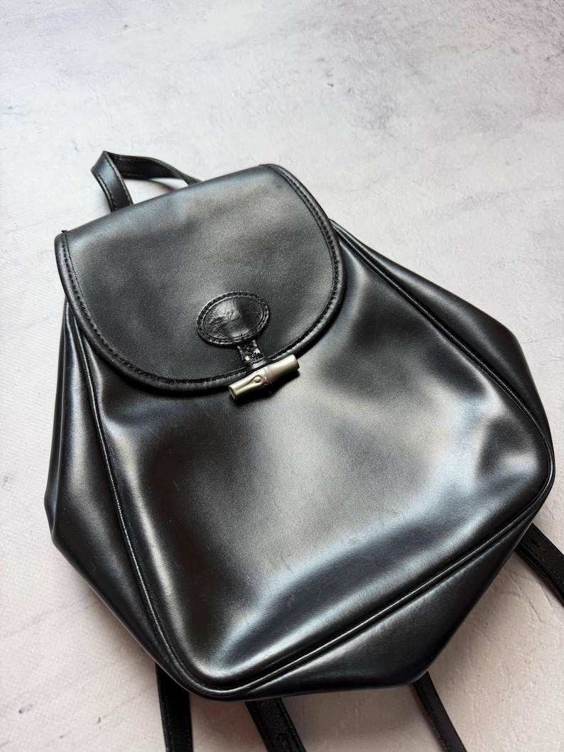 May include: A black leather backpack with a flap closure and a silver-tone clasp. The bag has a top handle and adjustable shoulder straps. The leather has a glossy finish, and the bag appears to be in good condition. The overall design is simple and classic.