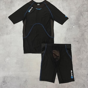 Puede incluir: Conjunto de camiseta y pantalón corto de compresión atlética negros. La camiseta de manga corta tiene costuras azules y el nombre de la marca "SKINS" en azul. El pantalón corto tiene un panel marrón y costuras azules.