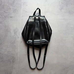 May include: A black leather backpack with adjustable shoulder straps and a top handle. The bag has a geometric shape and a glossy finish, set against a gray background. The straps are attached with silver-toned hardware.