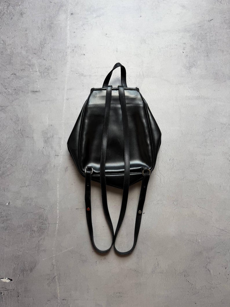 May include: A black leather backpack with adjustable shoulder straps and a top handle. The bag has a geometric shape and a glossy finish, set against a gray background. The straps are attached with silver-toned hardware.