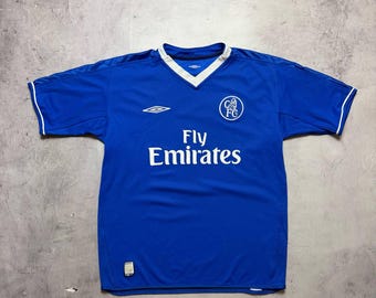 May include: A blue football shirt with white detailing on the collar and sleeves. The shirt displays the text "Fly Emirates" in white, a team emblem, and the Umbro logo. The jersey is made of a lightweight fabric.
