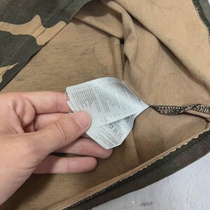 Puede incluir: Primer plano de una camiseta con estampado de camuflaje en tonos verdes, marrones y beige. Se ve una etiqueta con instrucciones de cuidado.