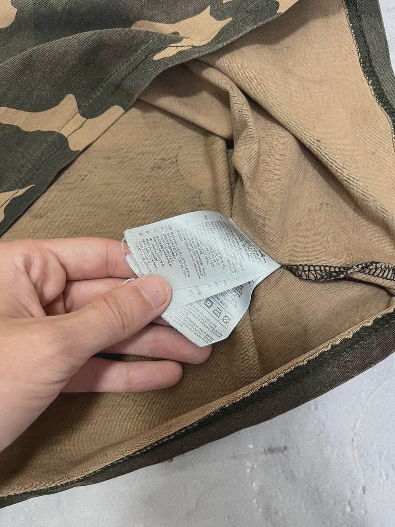 Puede incluir: Primer plano de una camiseta con estampado de camuflaje en tonos verdes, marrones y beige. Se ve una etiqueta con instrucciones de cuidado.