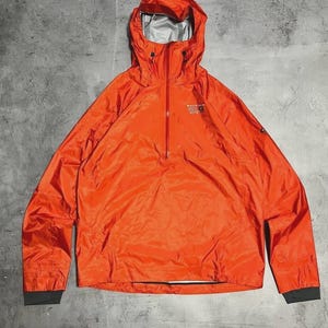 Puede incluir: Chaqueta anorak naranja brillante con capucha y media cremallera. La chaqueta tiene mangas largas con puños grises oscuros y un pequeño logotipo en el pecho. Fabricada con un material ligero, posiblemente resistente al agua.