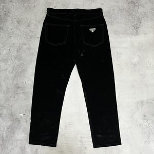 Pode incluir: Calças pretas de veludo com corte reto. As calças têm dois bolsos traseiros e um pequeno logotipo prateado no bolso traseiro direito. O tecido tem um ligeiro brilho, sobre um fundo cinzento.