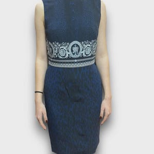 May include: Sleeveless navy blue dress with a decorative white and silver band at the waist. The dress has a subtle pattern and a knee-length hemline. The band features a crown and floral design.
