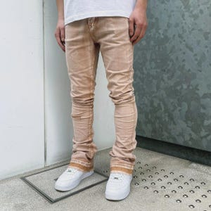May include: Tan denim jeans with a faded wash. The jeans have a slim fit and a slight flare at the ankles. The jeans are worn with white trainers. The model is wearing a white t-shirt.