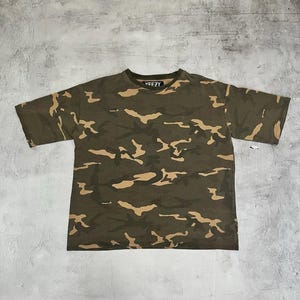 Puede incluir: Camiseta de camuflaje verde oliva con cuello redondo y mangas cortas. La camiseta presenta un estampado de formas marrones y beige. La palabra "YEEZY" est&aacute; impresa en blanco en el cuello.